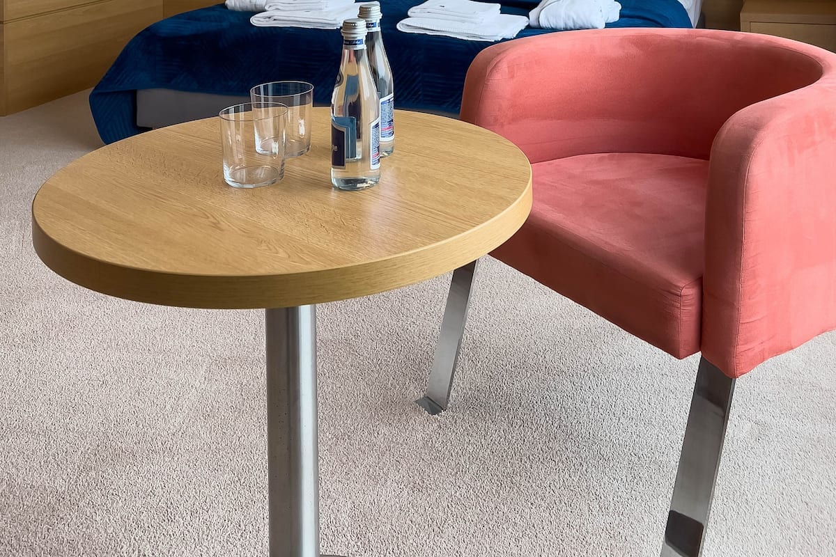 Ein Hotelzimmer mit einem Bett, einem runden Tisch mit Gläsern und Flaschen sowie einem rosa Sessel.
