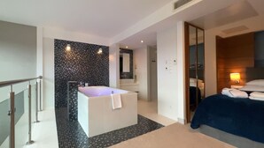 Een moderne hotelkamer met een groot ligbad, een aparte douche en een bed met een blauwe deken, plus een zithoekje met een lamp.