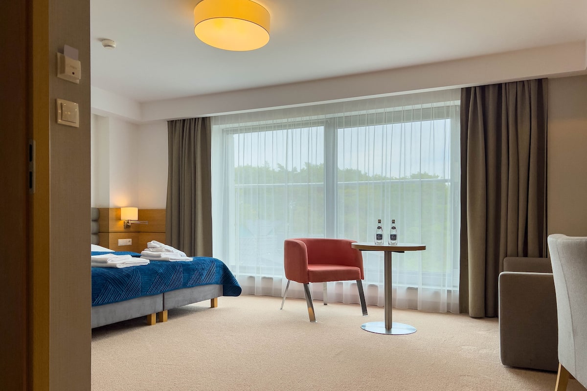 Ein Hotelzimmer mit einem Bett, einem roten Sessel, einem kleinen Tisch mit Flaschen und einem großen Fenster mit Vorhängen.