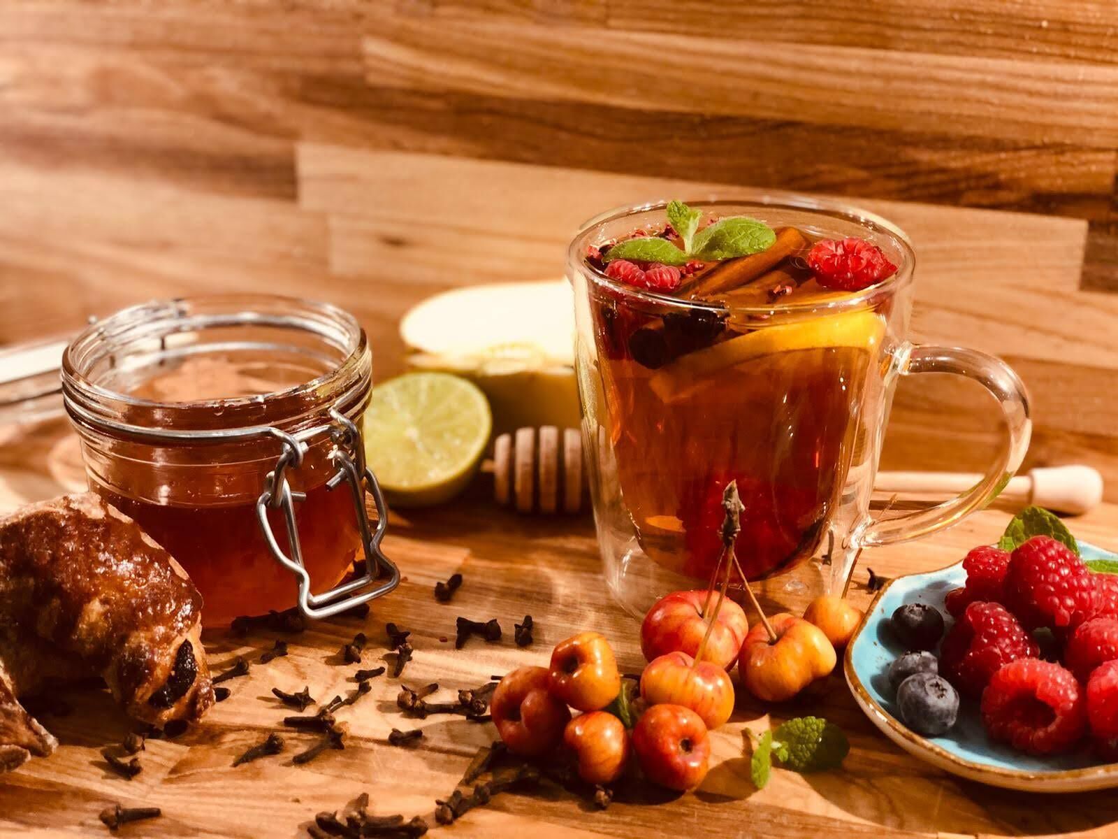 A glass jar of honey, a glass mug with tea, and a plate of berries on a wooden surface.