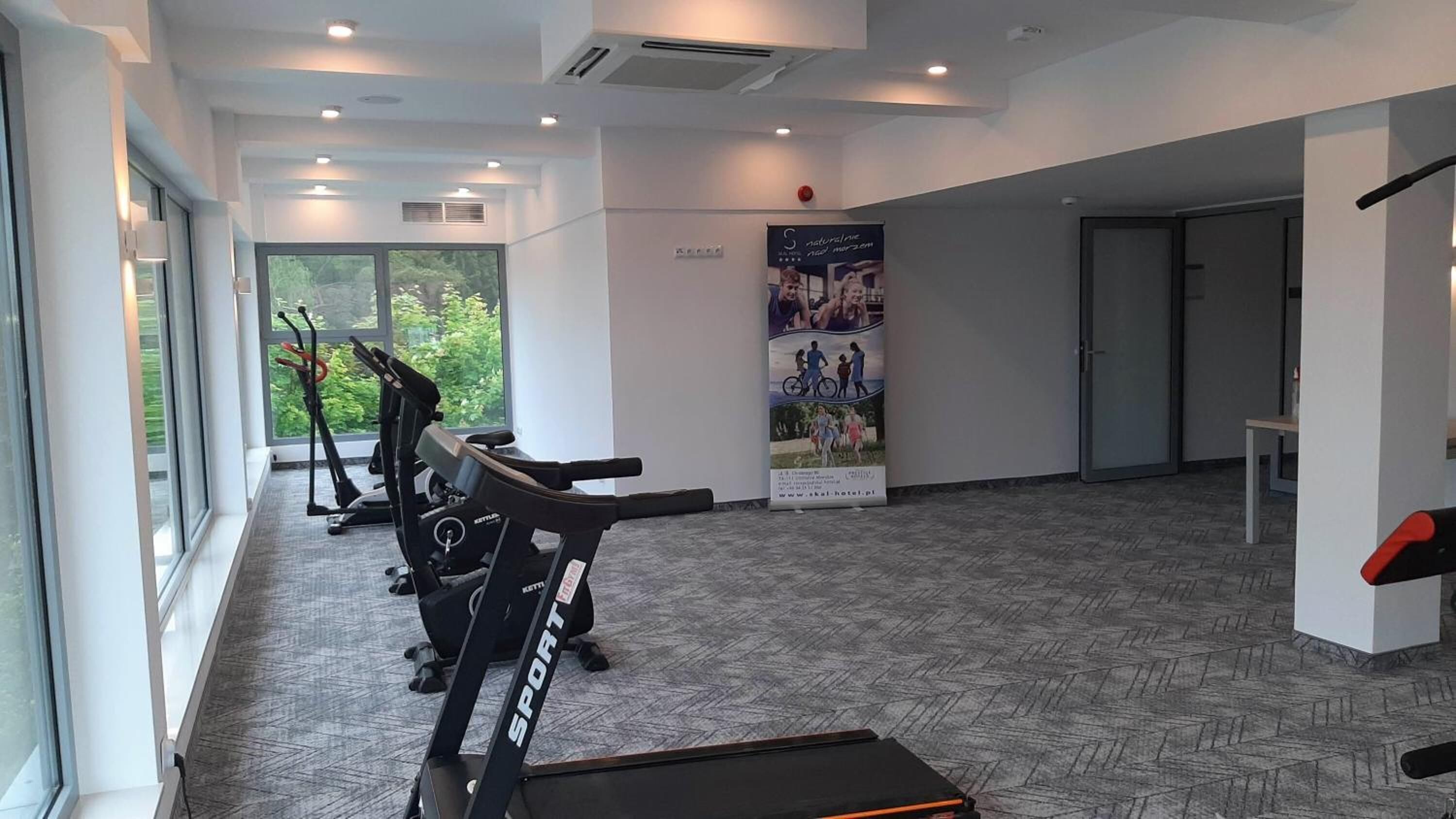 A gym with treadmills and stationary bikes, large windows, and a poster on the wall.