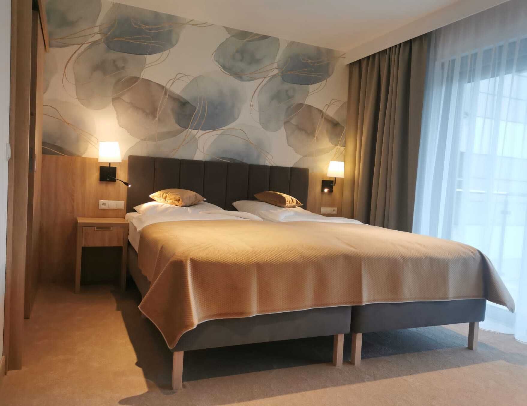 A hotel room with a large bed, two bedside tables, a wall mural, and a window with curtains.