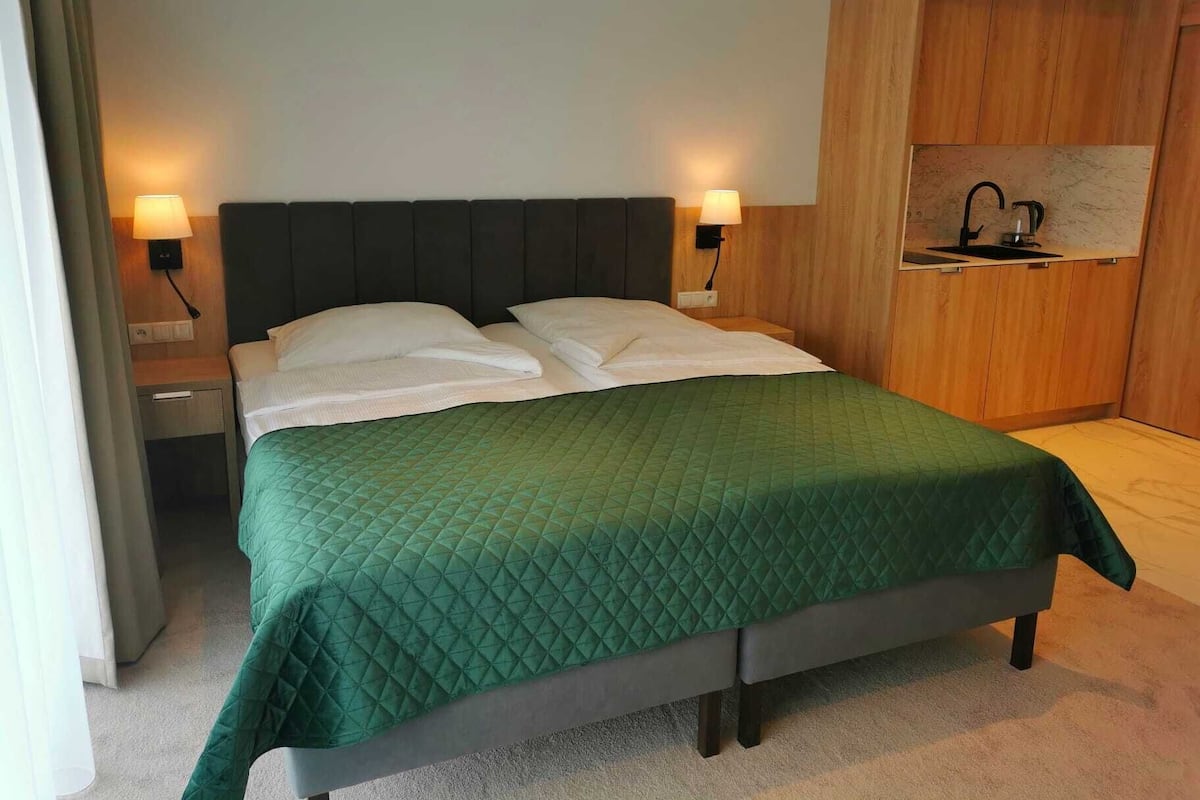 Ein Doppelbett mit einer grünen gesteppten Bettdecke, zwei Nachttischlampen, ein Holzschrank und eine kleine Küchenzeile mit einem Wasserkocher.