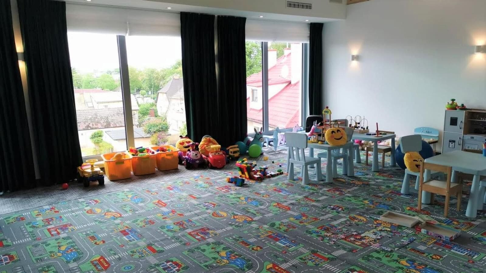Habitación amplia con ventanas grandes, zona de juegos con juguetes y un tapete de colores, además de un comedor con mesa y sillas.