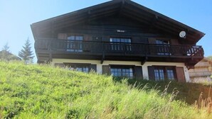 Un chalet à deux étages doté d’un balcon au niveau supérieur, entouré de verdure.