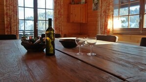 Une table de salle à manger en bois, avec une bouteille et deux verres à vin, un bol, et une fenêtre avec des rideaux.
