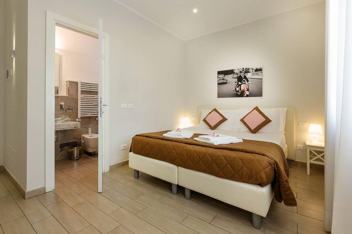 Un dormitorio con cama, una mesita de noche, un cuadro colgado en la pared y una puerta abierta que da a un baño.
