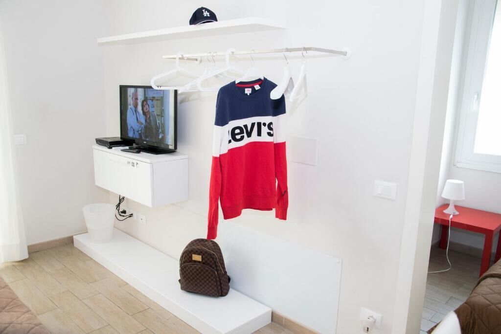 Una stanza con una scrivania bianca, un televisore, una camicia appesa e uno zaino.