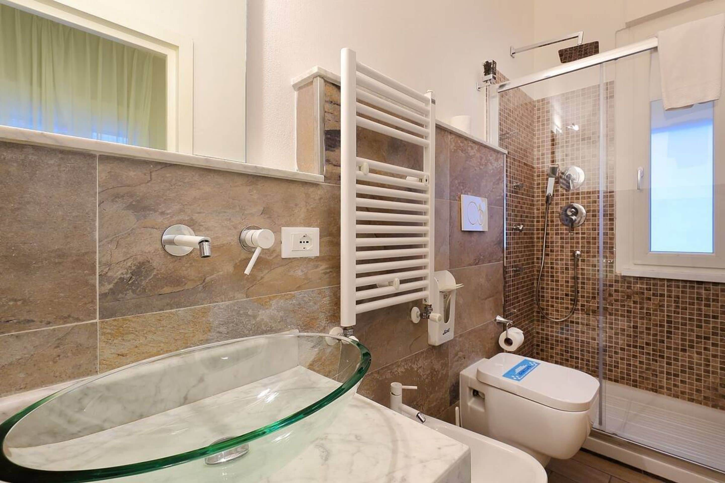 Un baño moderno con un lavamanos de vidrio, un inodoro blanco, una ducha con mampara de vidrio y un perchero para toallas.