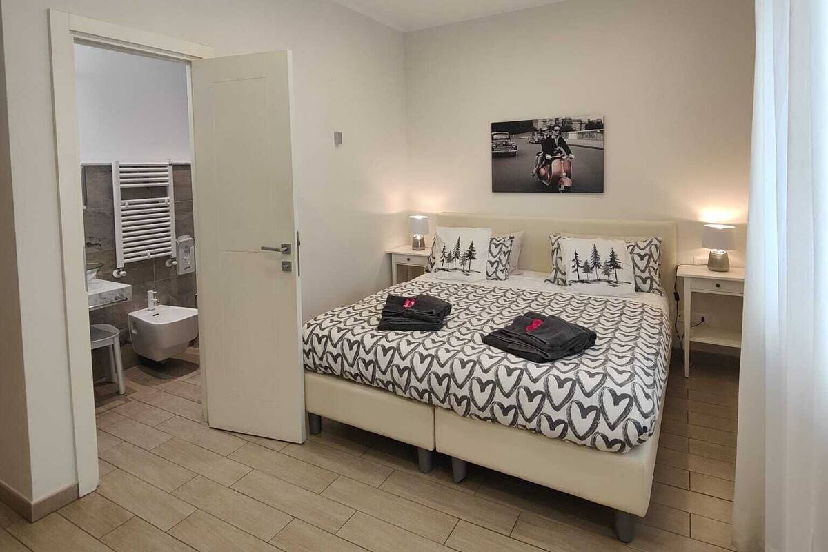 Un dormitorio con cama, mesitas de noche, un tocador, un baño con inodoro, lavabo y espejo, y una puerta que da a otra habitación.