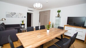 A modern living room with a wooden dining table, black chairs, a grey sofa, a flat-screen TV, and a potted plant.