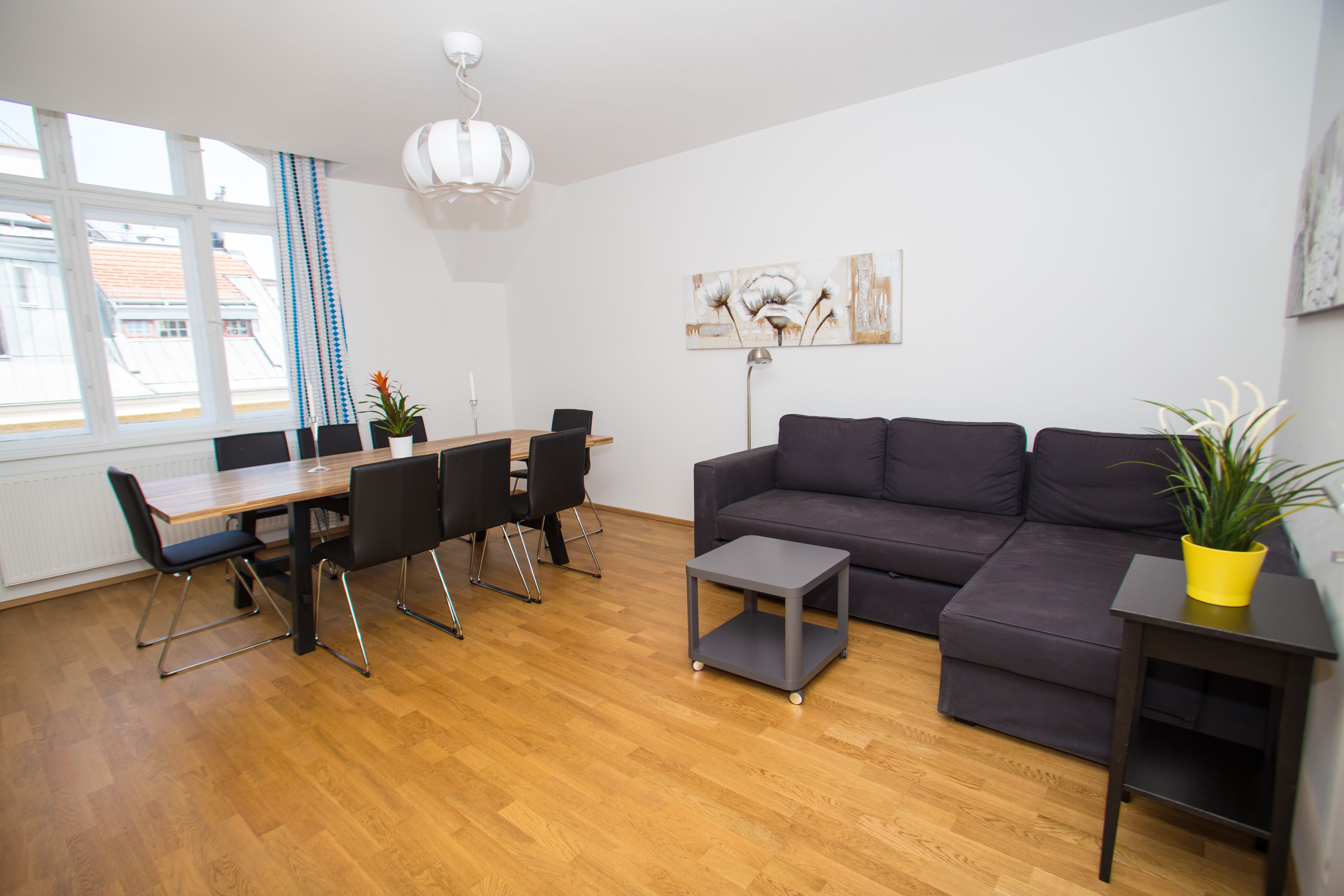 A modern living room with a wooden floor, a grey sofa, a dining table with chairs, and a small side table with a plant.