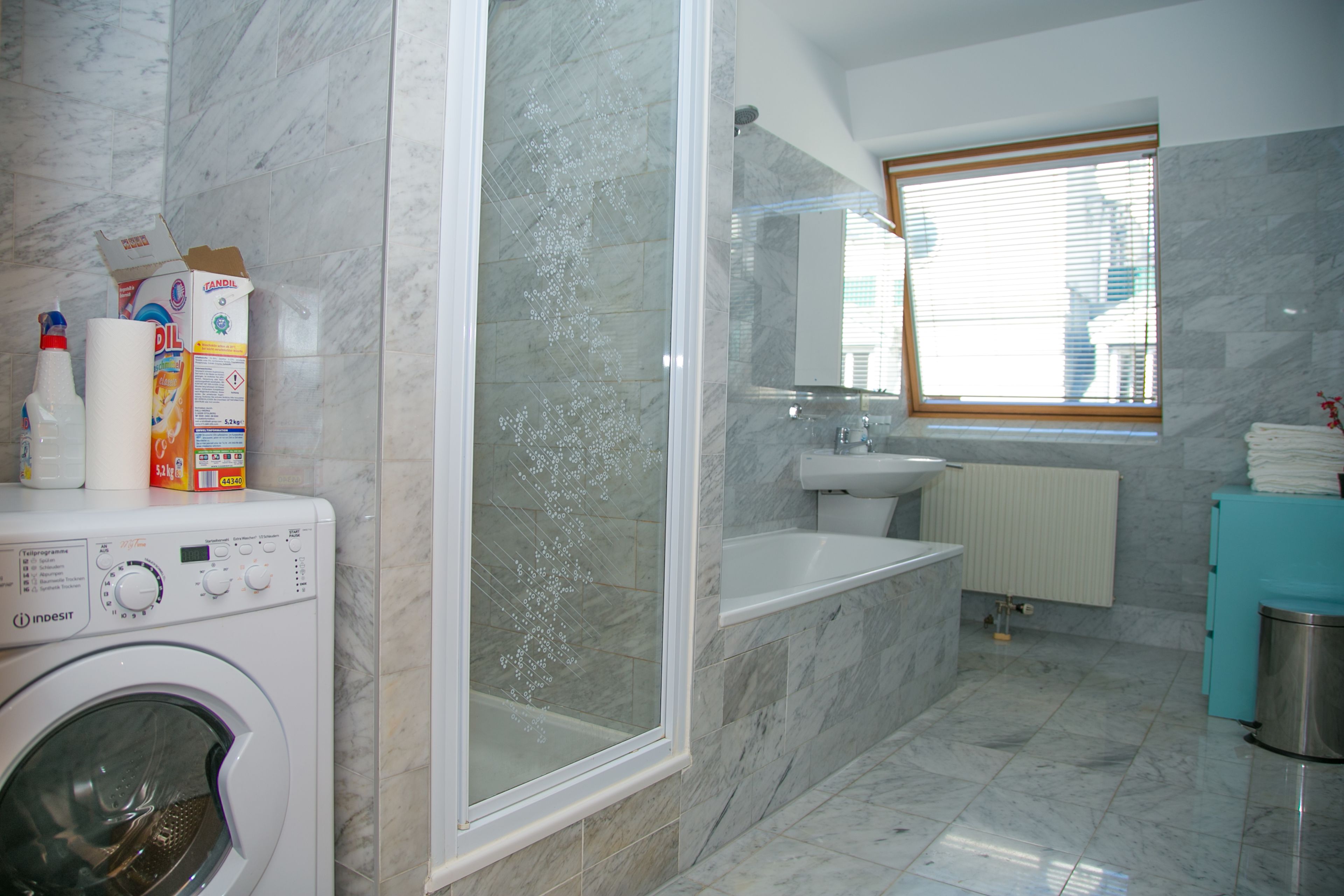 A modern bathroom with a washing machine, a glass-enclosed shower, a sink, and a window with blinds.