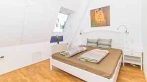 Una camera da letto con soffitto spiovente, un letto con lenzuola a righe, un comodino, una lampada a parete e una finestra con vista sugli edifici.