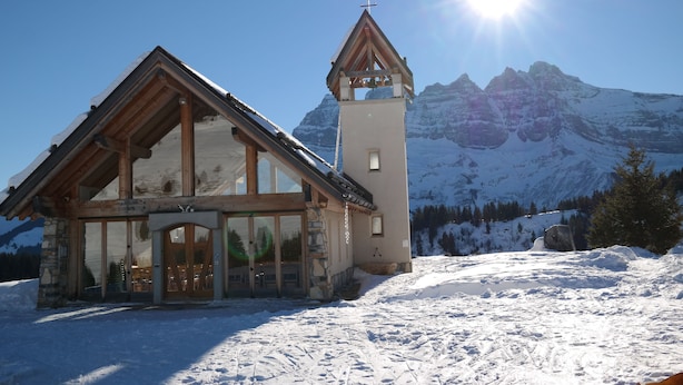 A wooden lodge with a bell tower and large windows, set against a snowy mountain backdrop.