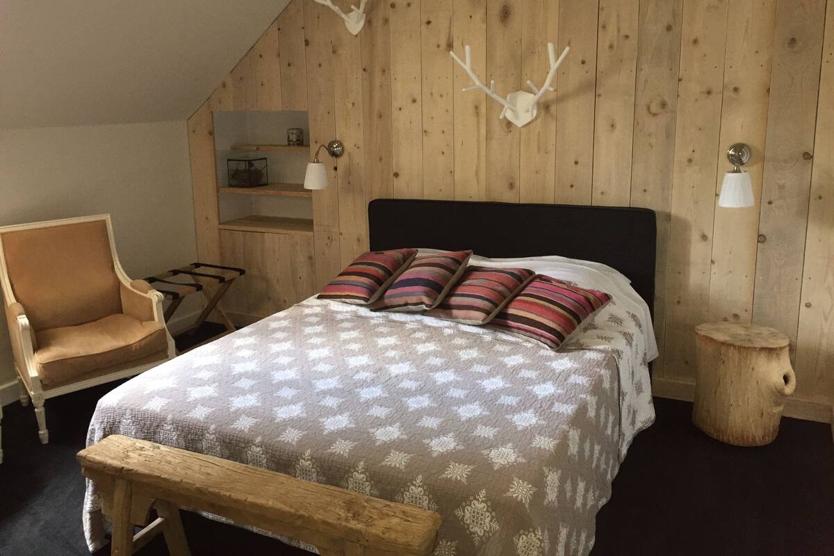 Quarto com teto inclinado, cama com roupa de cama estampada, uma cadeira, mesa de cabeceira de madeira e chifres fixados na parede.