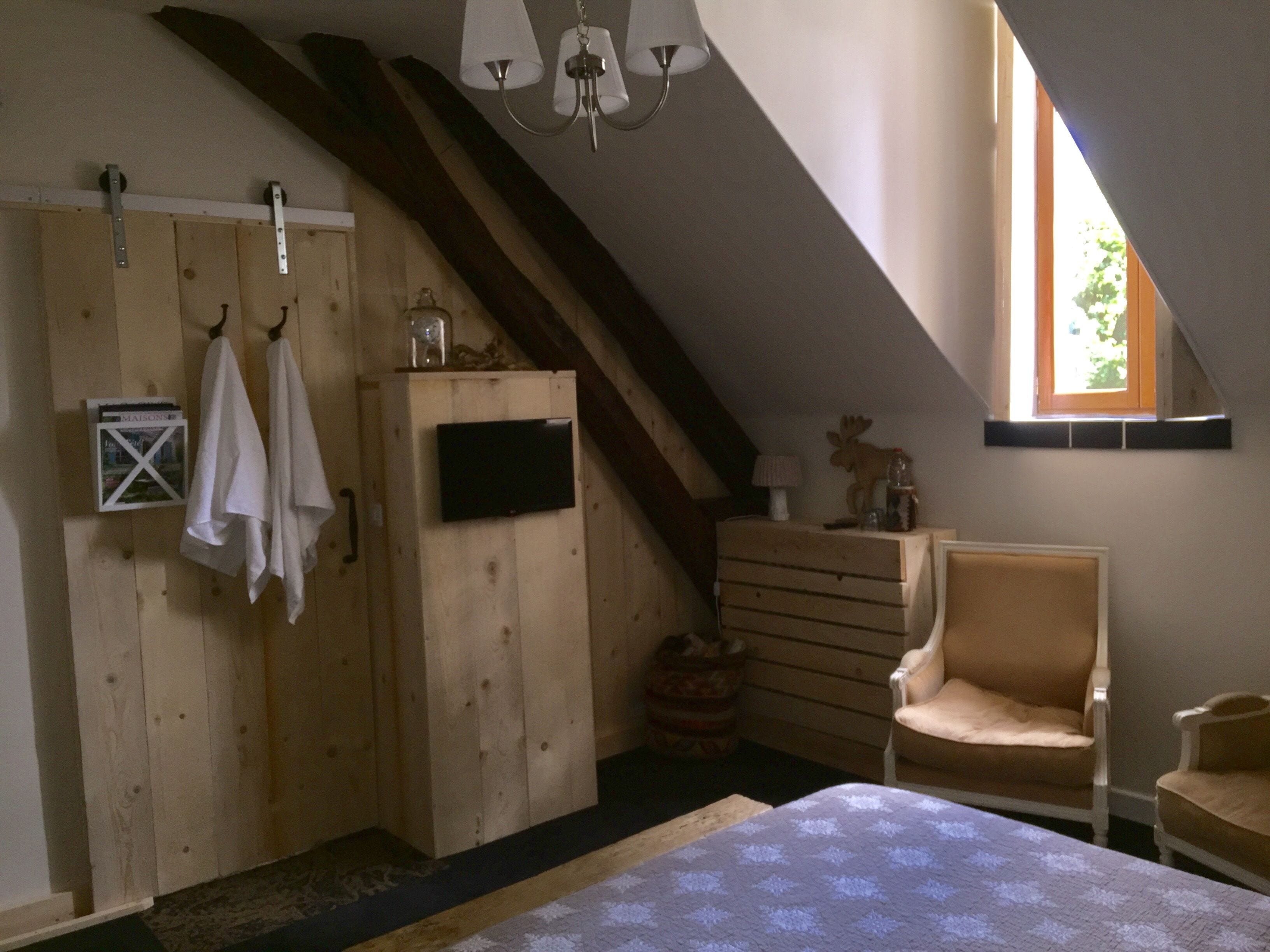 A bedroom with a sloped ceiling, wooden wardrobe, two chairs, a bed with a patterned bedspread, and a window with natural light.