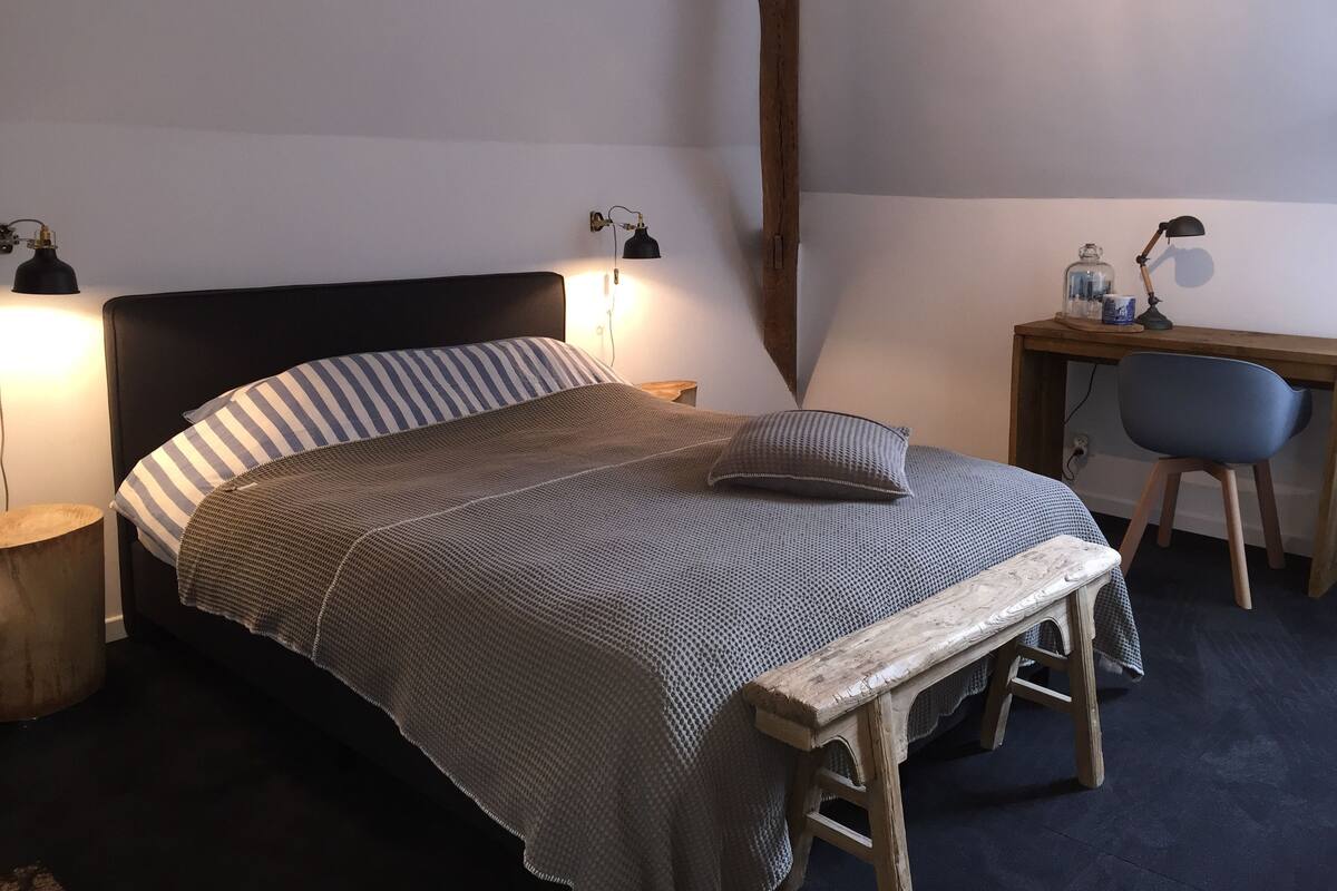 Quarto com cama, luminárias de cabeceira, escrivaninha de madeira, cadeira e pequena mesa com abajur.