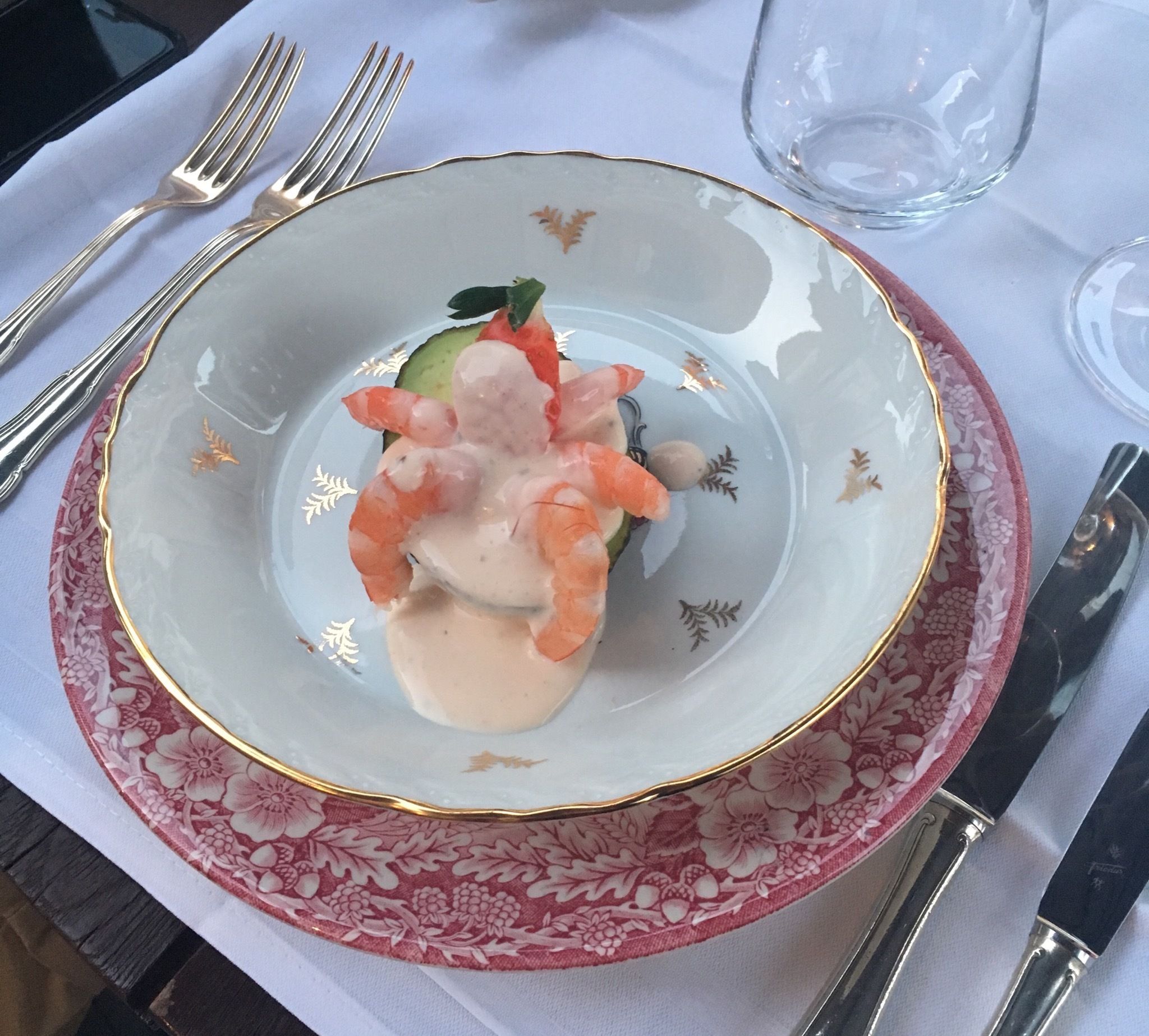 A plate of shrimp with sauce, a cucumber, and a small green vegetable on a table with a floral-patterned plate and a glass of water.