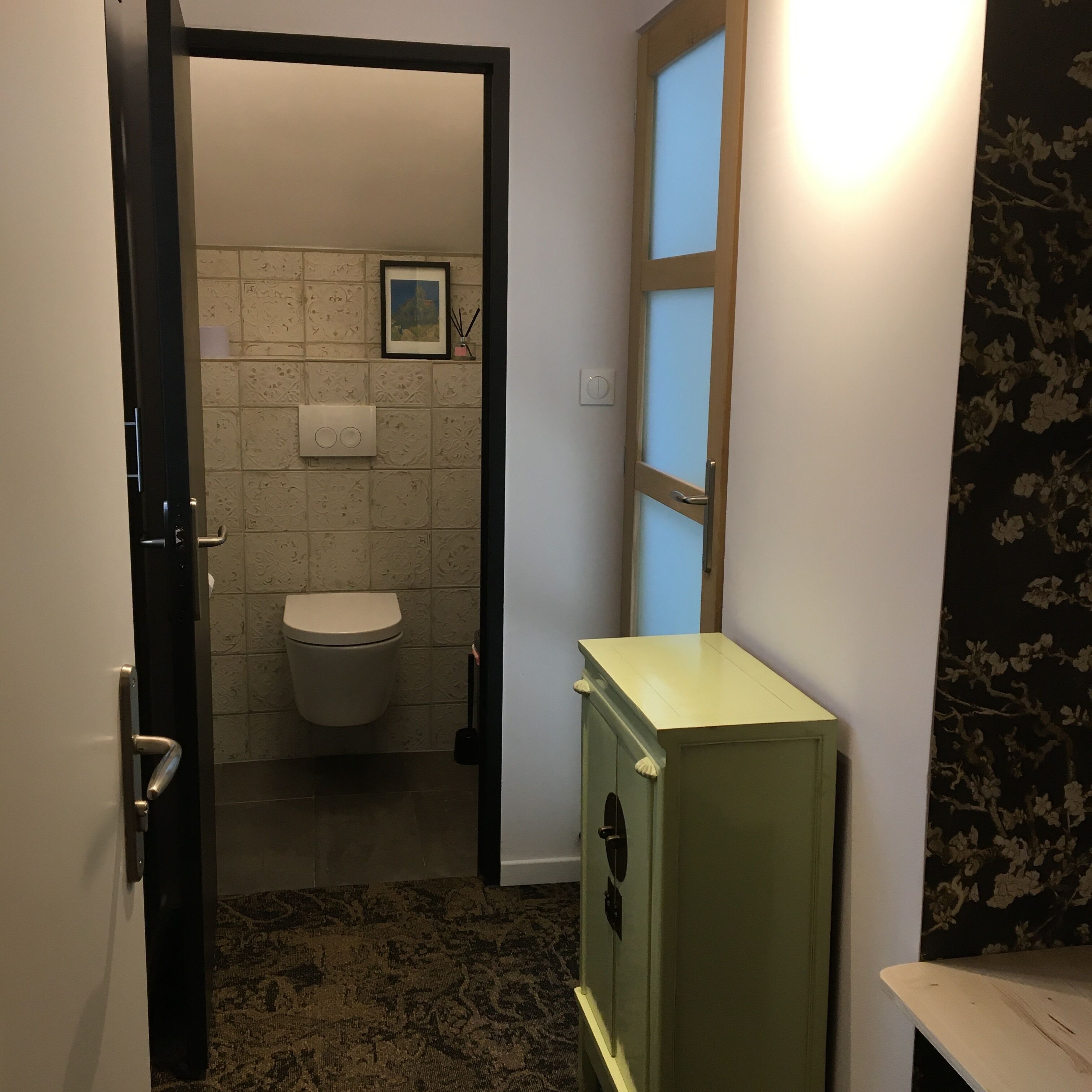 Une salle de bain avec des toilettes, un luminaire fixé au mur et un petit meuble.