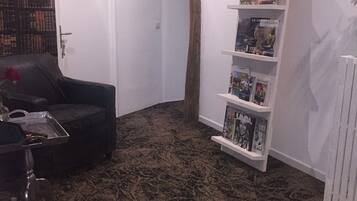 A living room with a dark patterned carpet, a black sofa, a small coffee table, and a bookshelf with magazines.