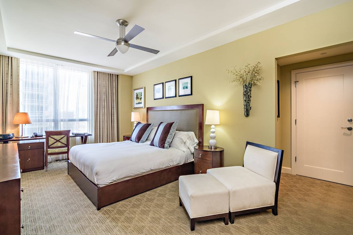A hotel room with a bed, bedside tables, a desk, a chair, a ceiling fan, and a door.