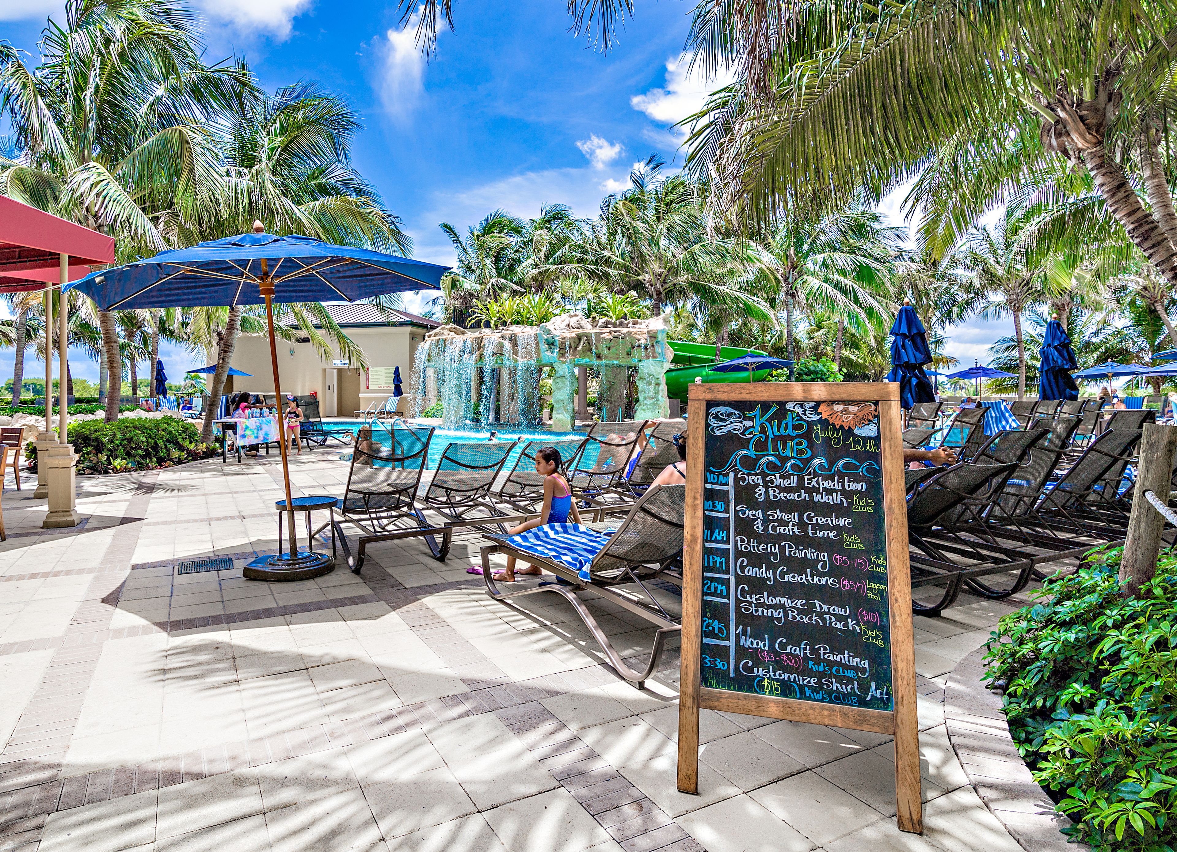 A poolside area with lounge chairs, umbrellas, and a chalkboard listing activities.