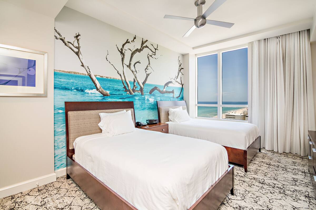 A hotel room with two beds, a large window with a view of the sea, a ceiling fan, and a mural of a tree and water.