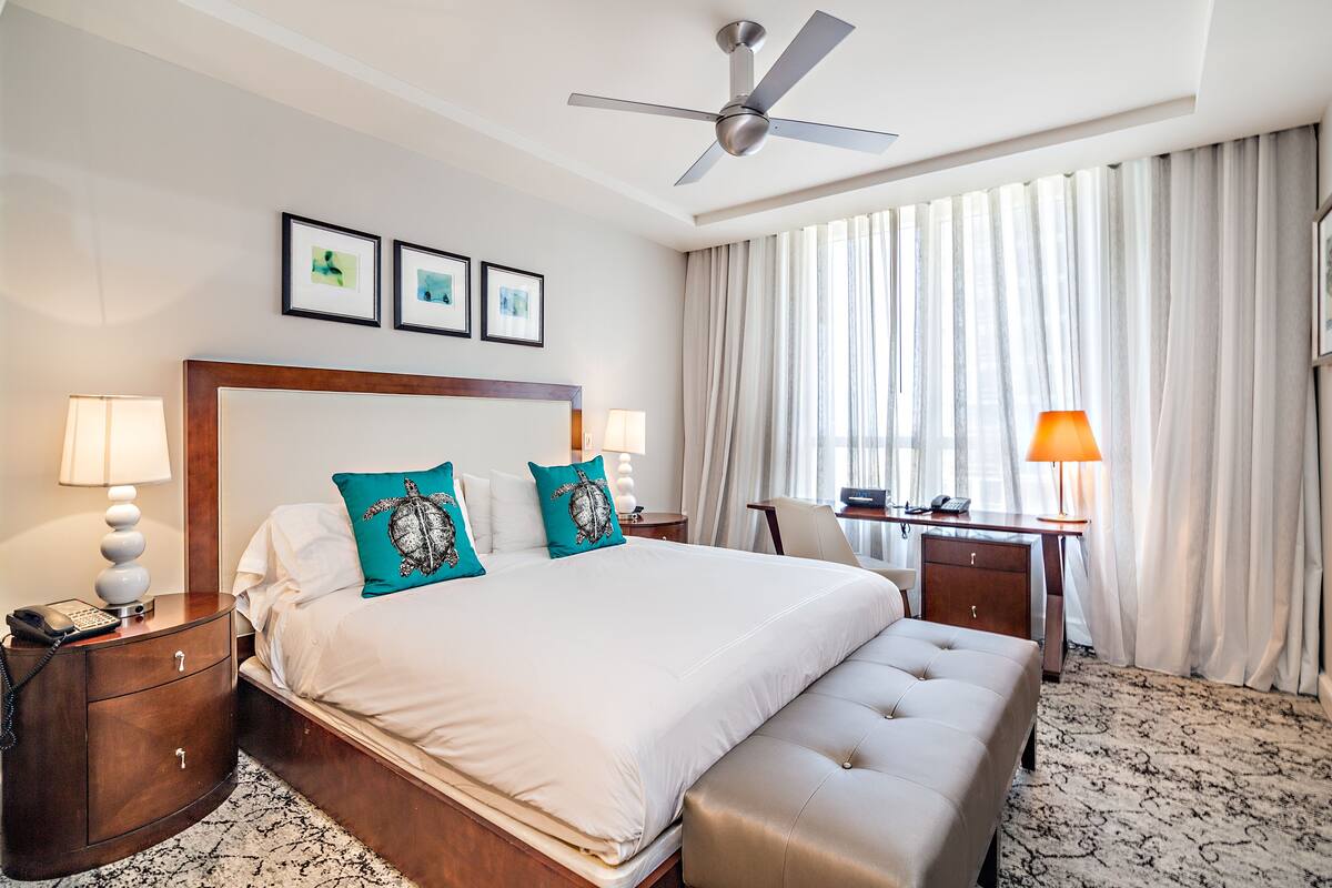 A hotel room with a large bed, a desk, a chair, a ceiling fan, and a window with curtains.