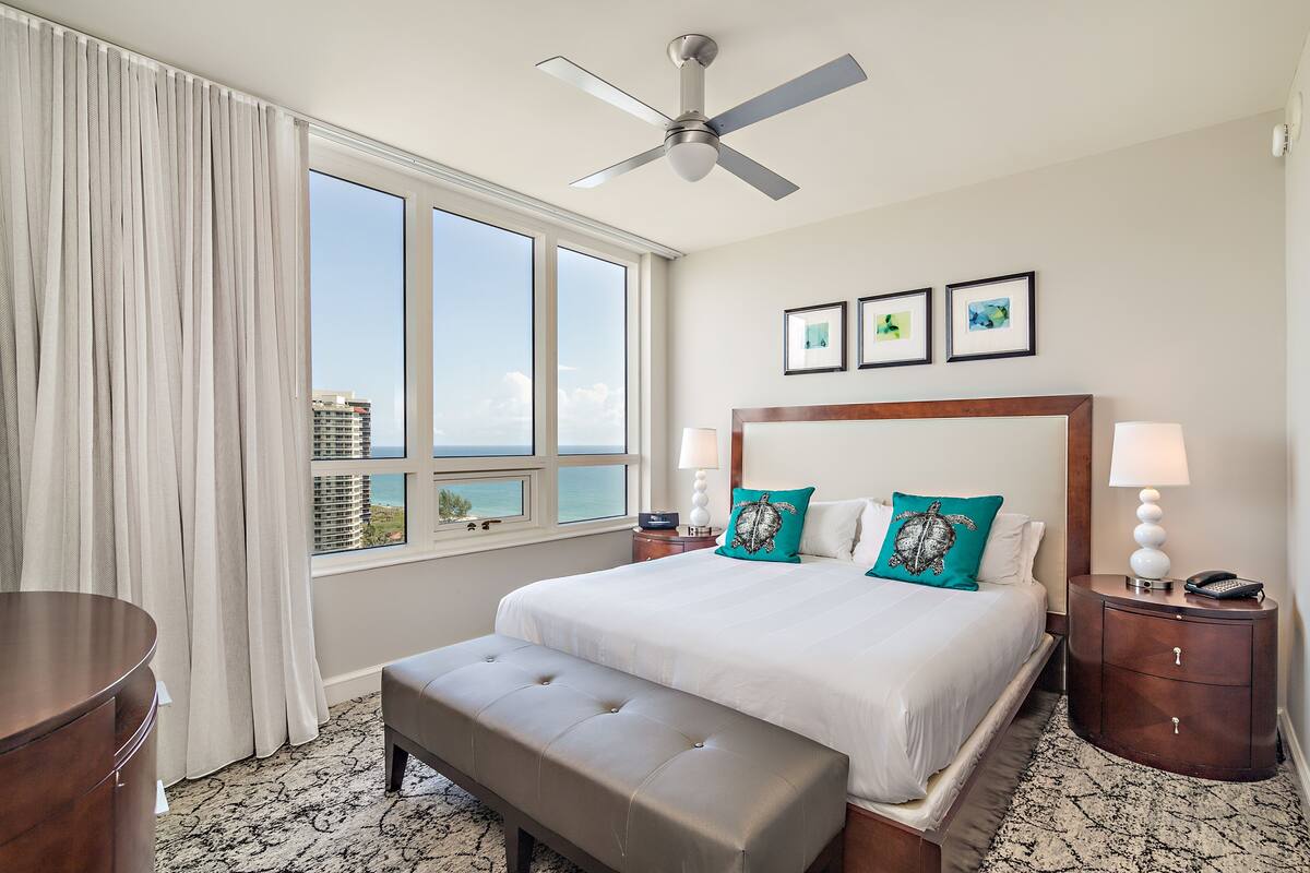 A hotel room with a large bed, a ceiling fan, a view of the city, and a desk with a lamp.