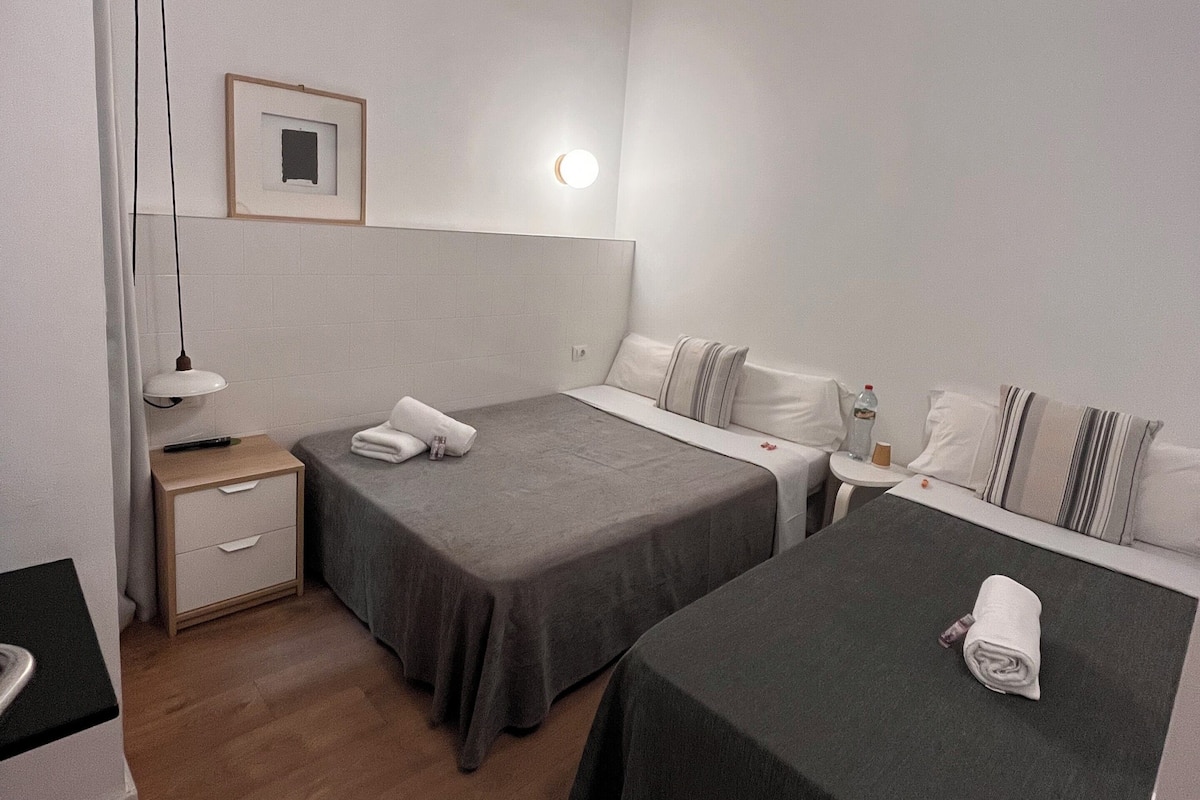 Une chambre d’hôtel avec deux lits simples, une table de chevet avec une lampe, une petite table de chevet avec une bouteille, et un tableau encadré au mur.
