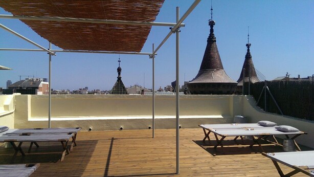 A rooftop terrace with wooden tables and benches, a pergola, and two pointed towers in the background. - Hostal Argo (Barcelona)