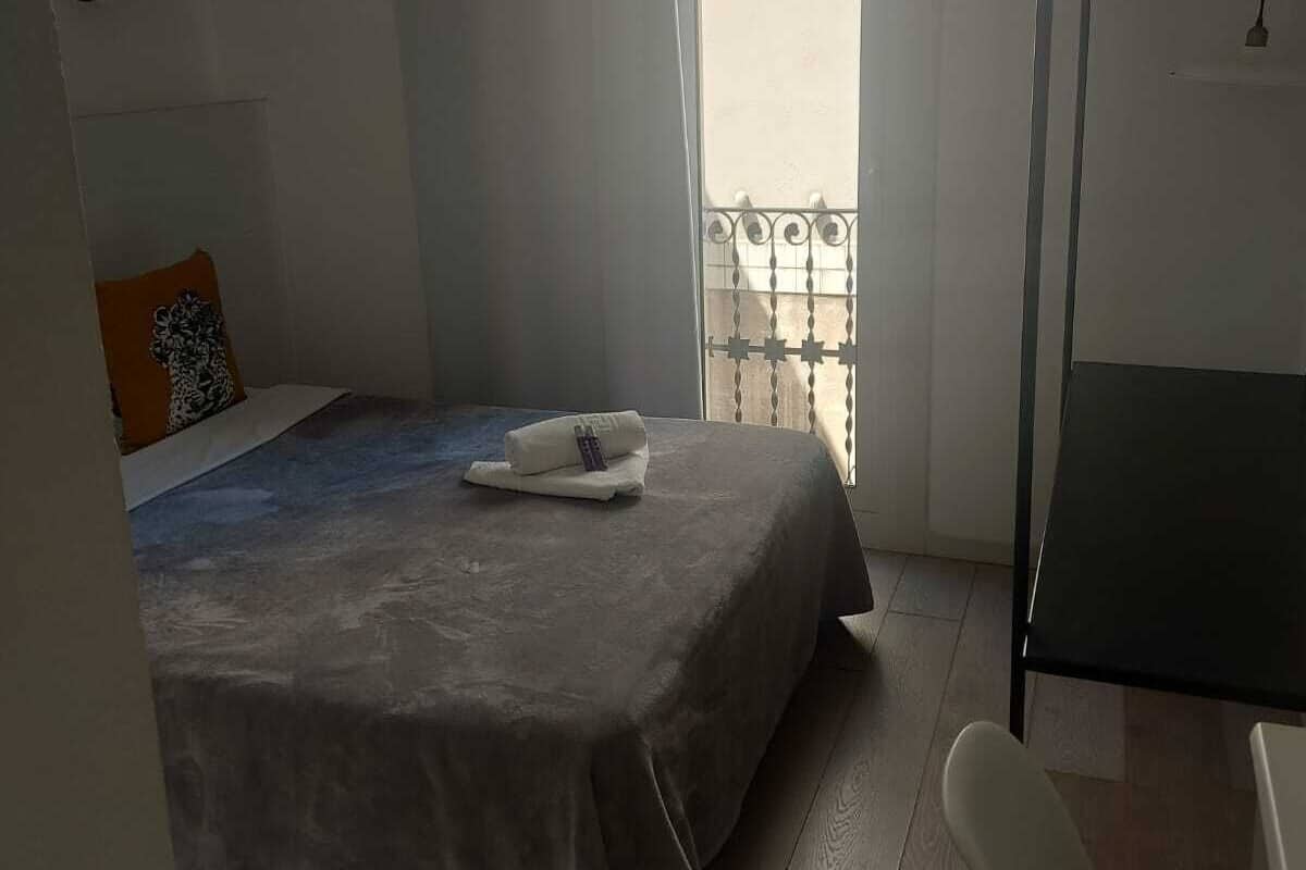 Une petite chambre à coucher, bien rangée, avec un lit, une chaise, une table et un balcon dot é d’une balustrade.