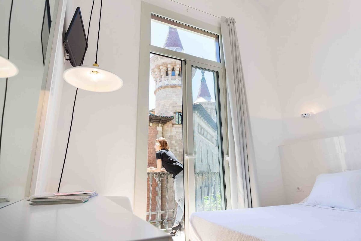 Une chambre d’hôtel avec une grande fenêtre, un lit, un bureau avec une lampe et une vue sur une tour.