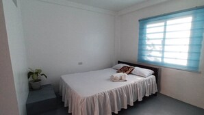A small, single-bed room with a plain white ceiling, a window with blue blinds, and a potted plant on a small ledge. - Old Parola Seaside Cottages (Baler)