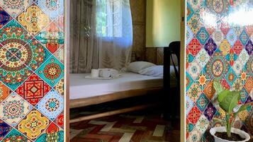 A room with a bed, a chair, a potted plant, and a window with a sheer curtain.