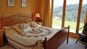 Een slaapkamer met houten vloer, een bed, nachtkastje, lamp en stoel. Twee ingelijste foto's hangen aan de muur en er is uitzicht op een bos door het raam.