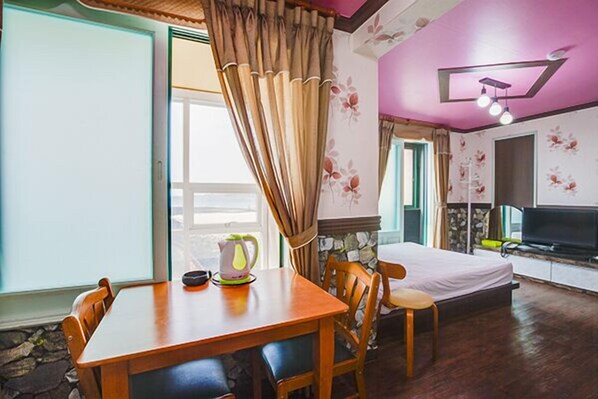 Huoneessa on puinen ruokapöytä, tuoleja, sänky, televisio ja kiviseinä.