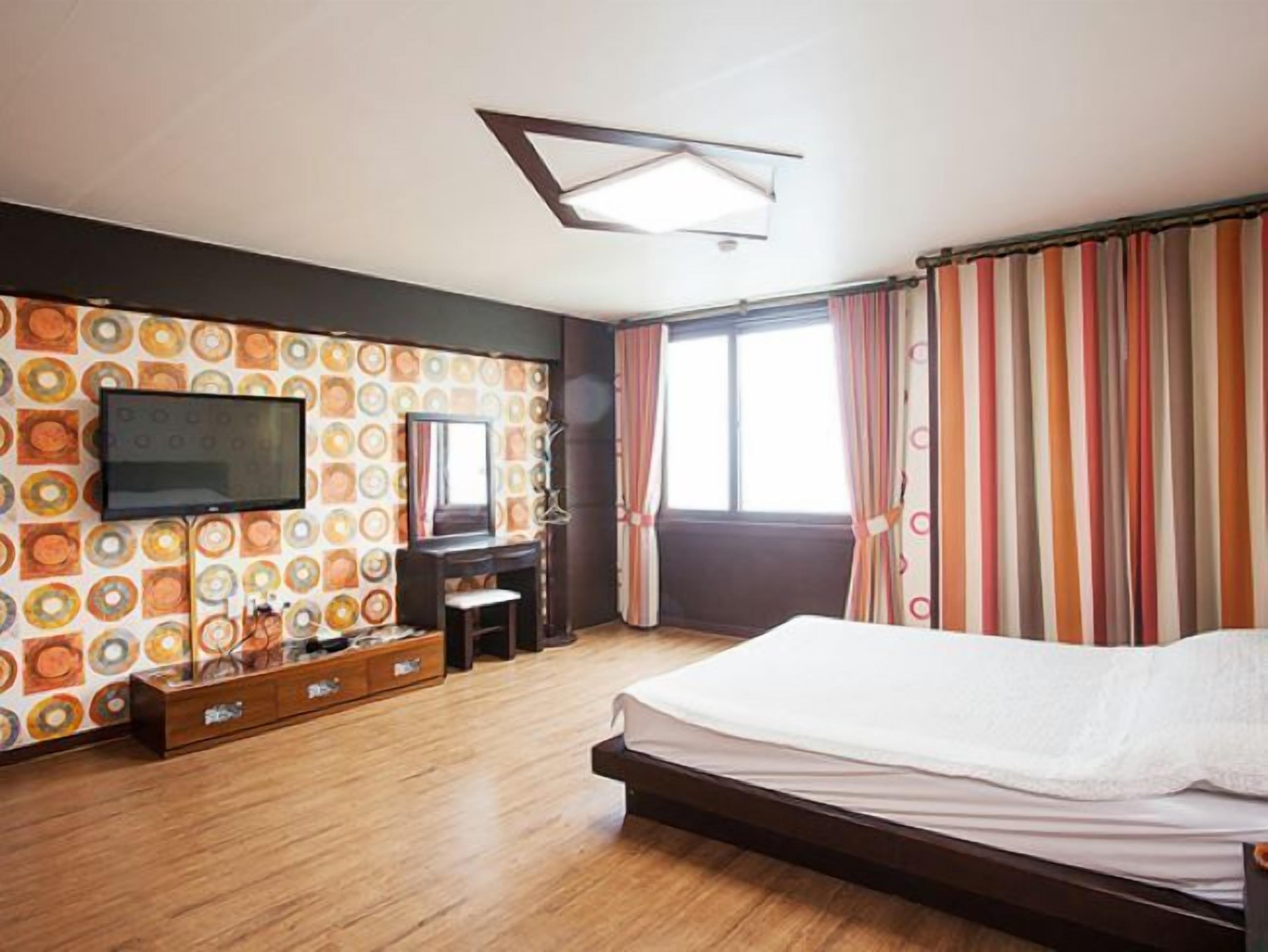 Quarto com cama, televisão na parede, penteadeira e janela com cortinas.