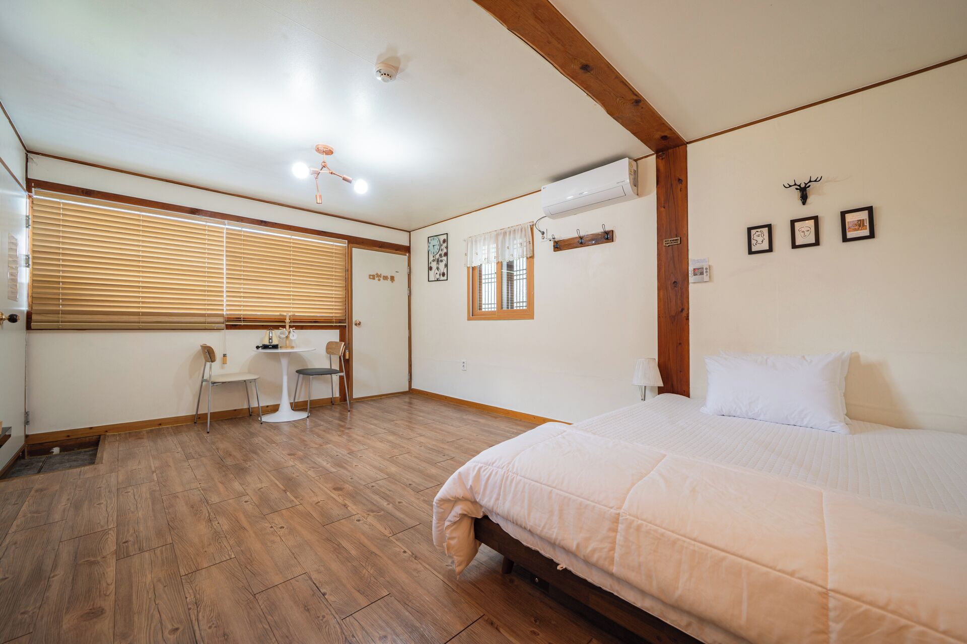 A bedroom with a bed, wooden flooring, a small table with chairs, a window with blinds, and a ceiling fan.