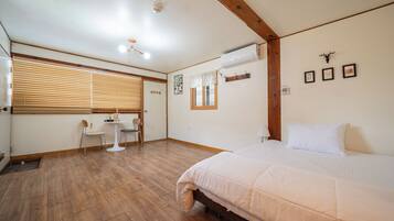 A bedroom with a bed, wooden flooring, a small table with chairs, a window with blinds, and a ceiling fan.