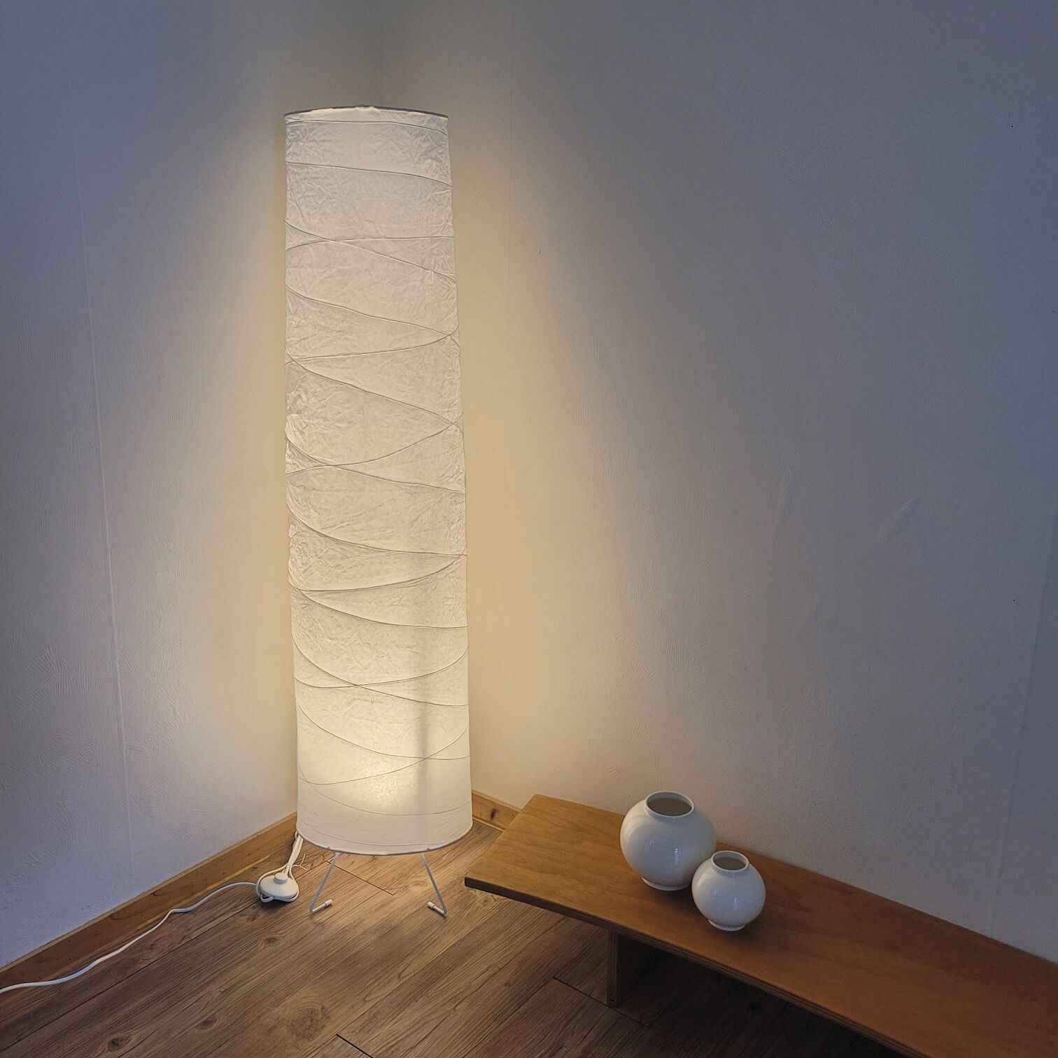 A cylindrical floor lamp with a textured surface, a wooden table with two white vases, and a power cord on the floor.