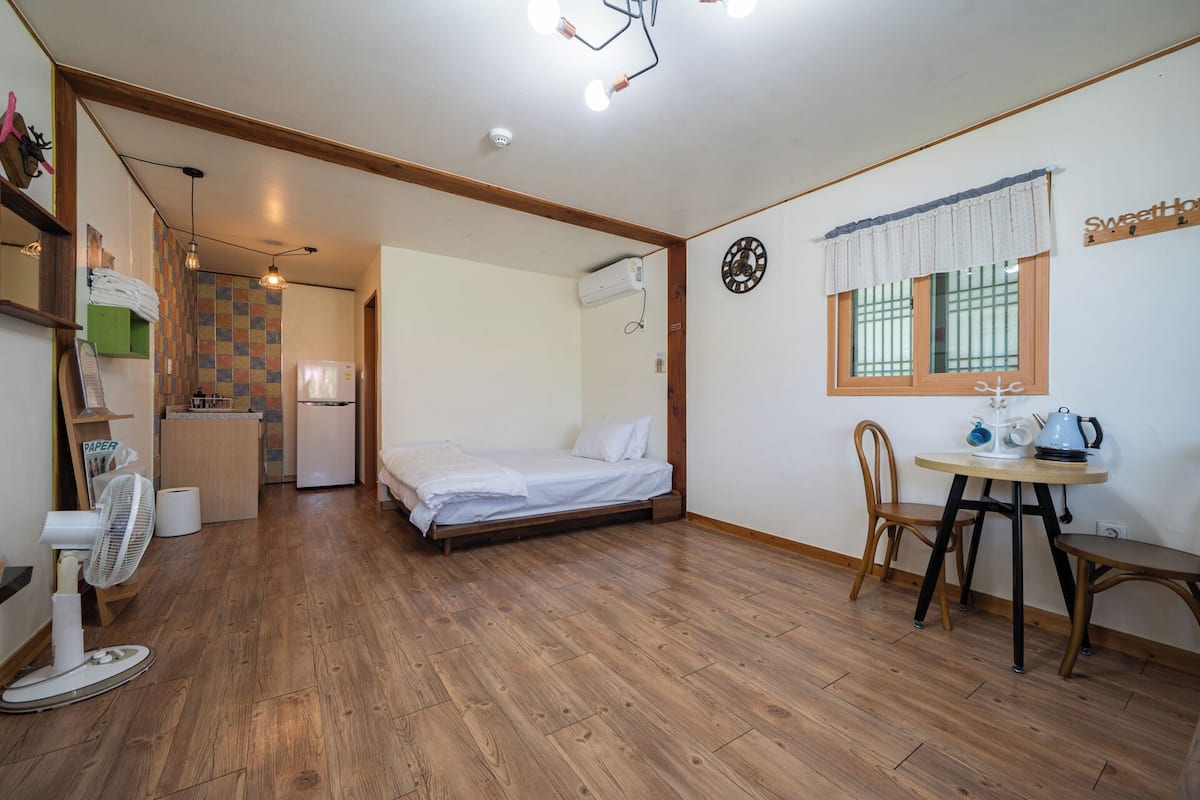 A compact room with a bed, a small table with a kettle, a fan, and a kitchenette area with a refrigerator and a sink.