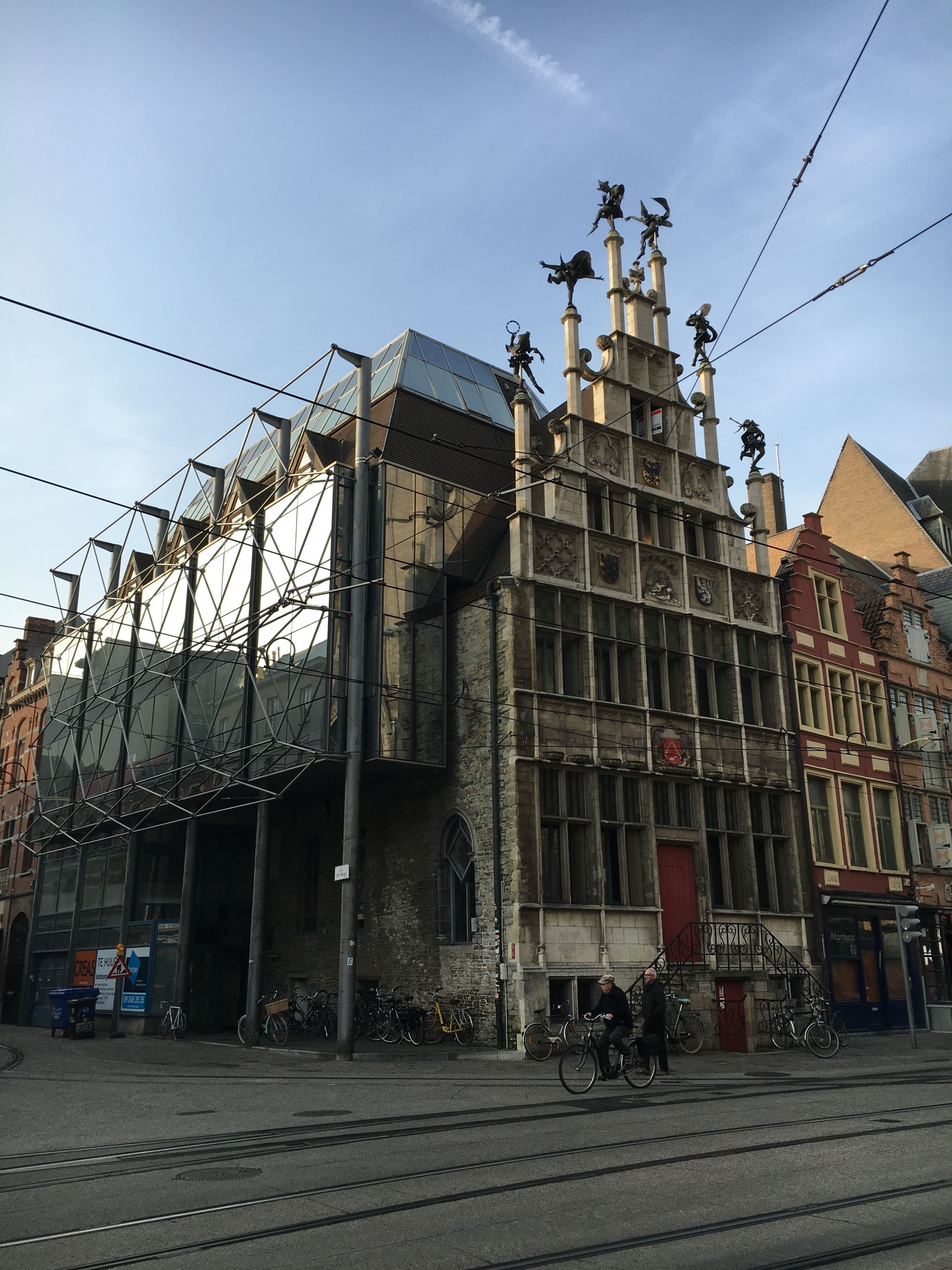 Een historisch gebouw met een moderne glazen uitbouw, met een rode deur en meerdere beelden op de gevel.
