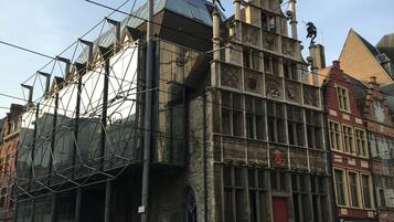 Een historisch gebouw met een moderne glazen uitbouw, met een rode deur en meerdere beelden op de gevel.