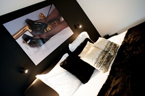 Un lit bien fait, avec une literie blanche et noire, un tableau encadré représentant des chaussures accroché au mur et une tête de lit de couleur sombre.