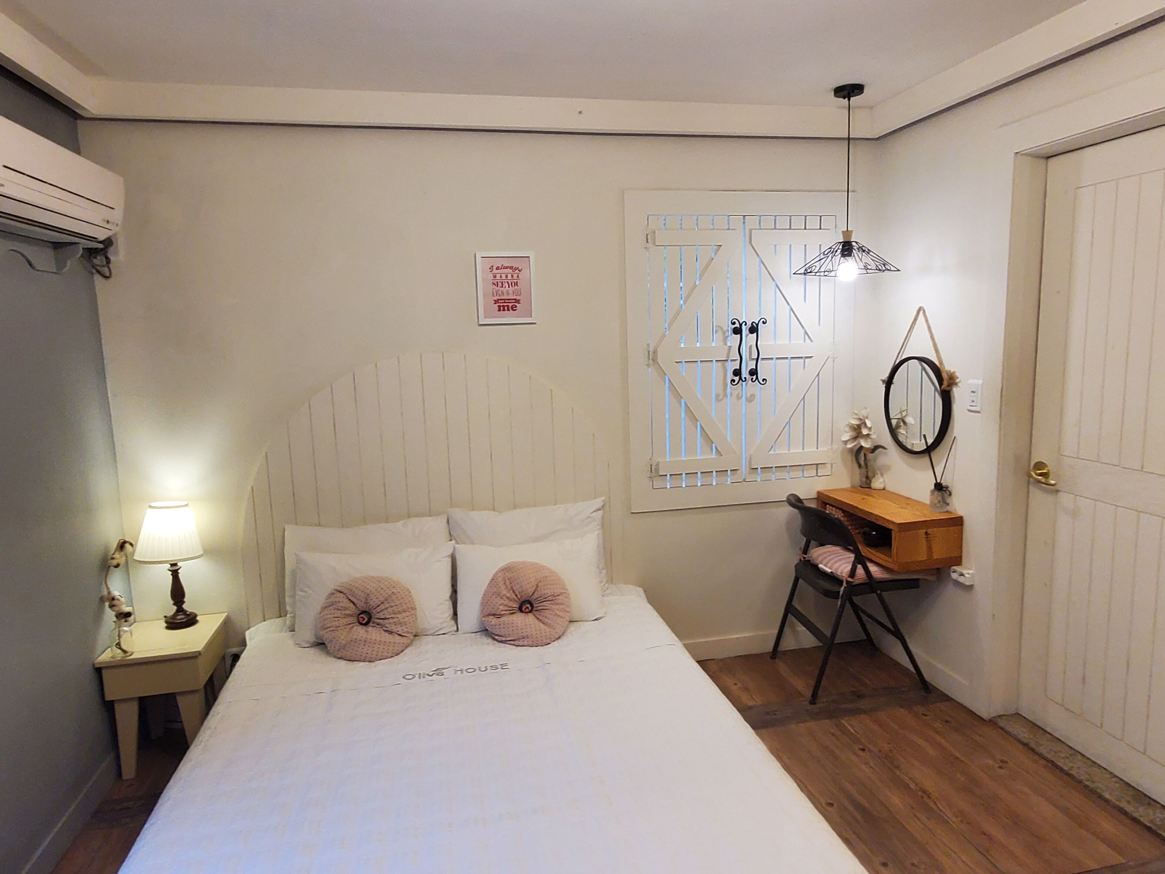 A bedroom with a bed, bedside table, lamp, window with blinds, and a dresser with a mirror.