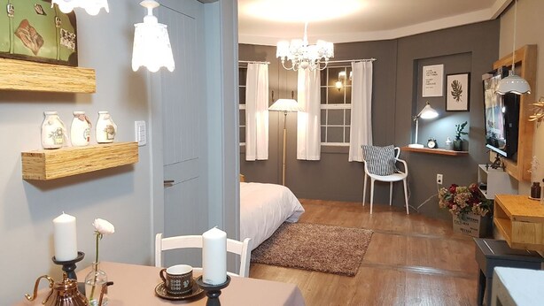 A cozy bedroom with a bed, bedside table, and a chair. There is a wooden shelf with decorative items and a small table with a candle and a teapot. - Gyeongju Olive House Pension (Gyeongju)