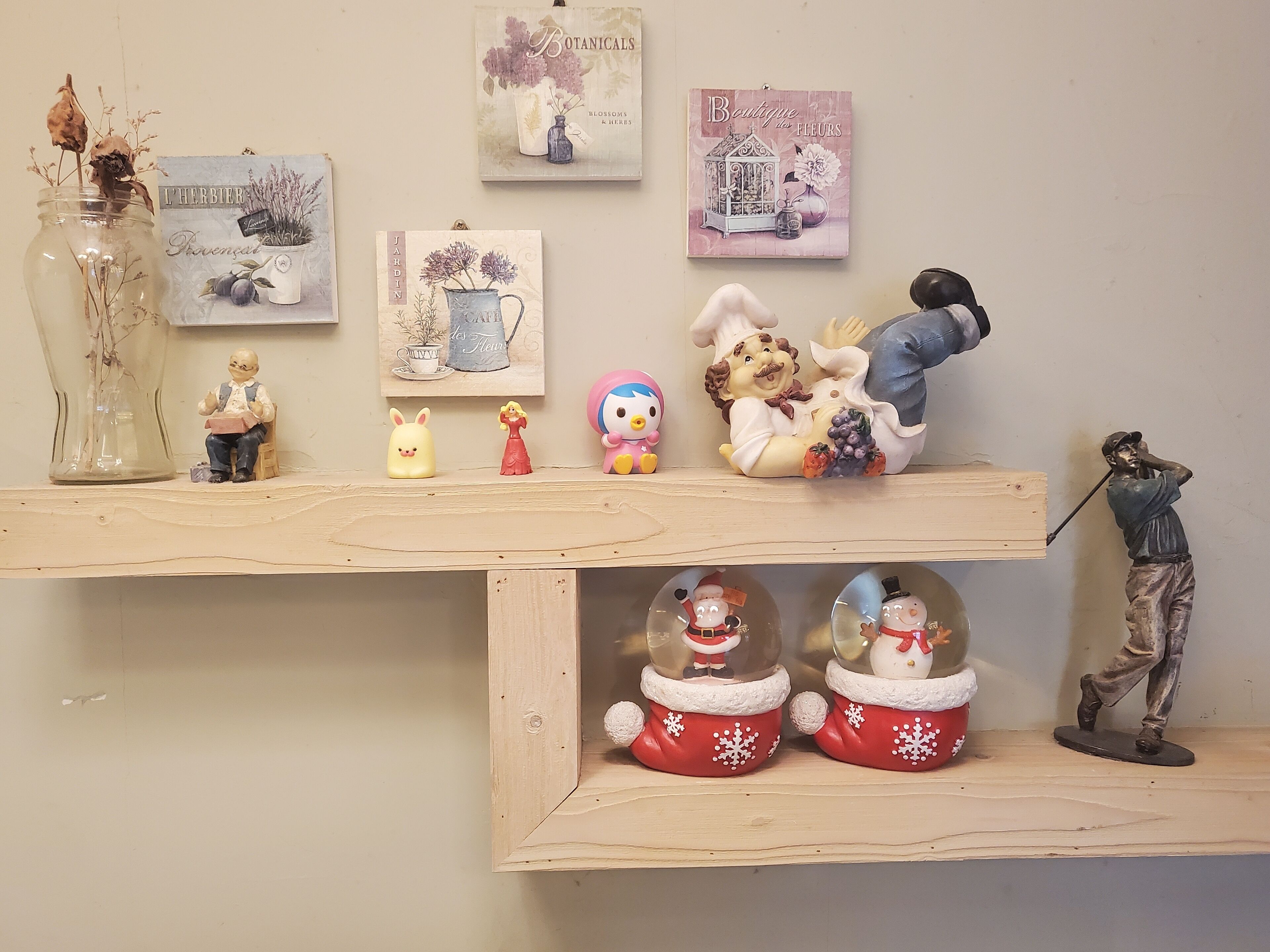 A wooden shelf with various decorative items including figurines, a vase with dried plants, and framed botanical prints.