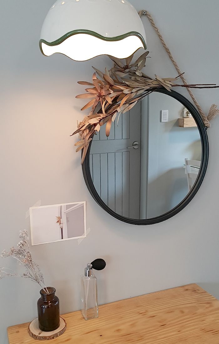 Et spejl med rund ramme, en vase med visne blade og en lampe på en træoverflade.